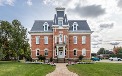 A two story brick house with slate grey roof, central second story balcony, and several symmetrical white trimmed windows is centred in the frame, surrounded by blue sky and green grass. 