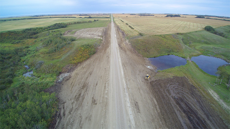 Vue aérienne d’une nouvelle route en gravier, entre des champs verts et dorés, avec une chargeuse de matériaux de construction près d’un petit marécage.