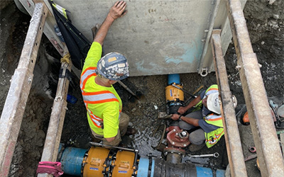 Two men in high visibility vests and hard hats stand in shallow water and silt, surrounded by concrete barriers and water main pipes. One worker stands, hand leaning on concrete barrier, looking down on kneeling colleague as he works on water main.