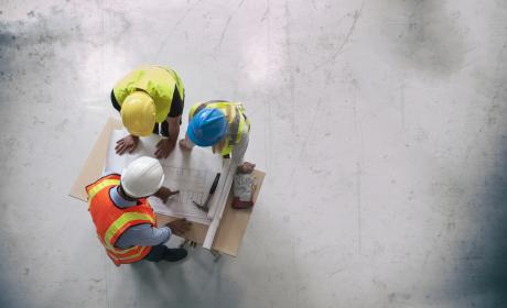 Group of three engineers in hardhats overlook blueprints in a new building. 