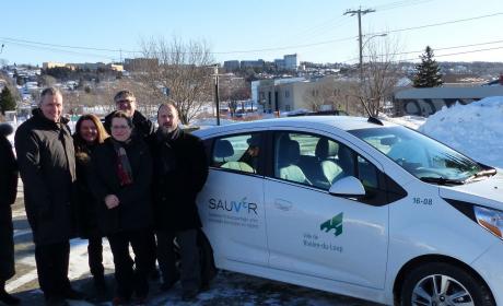 Des représentants des municipalités sourient à la caméra devant l’un des véhicules électriques, par une journée d’hiver ensoleillée.
