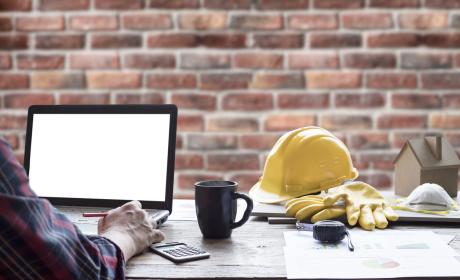 Engineer sitting at desk working on a laptop with his yellow hardhat beside him.